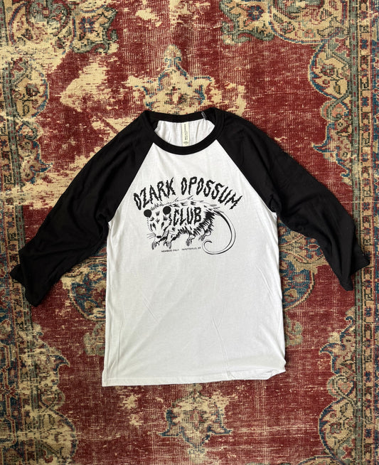 Black and white baseball shirt with 'Ozark Opossum Club' design on a textured red and brown rug.