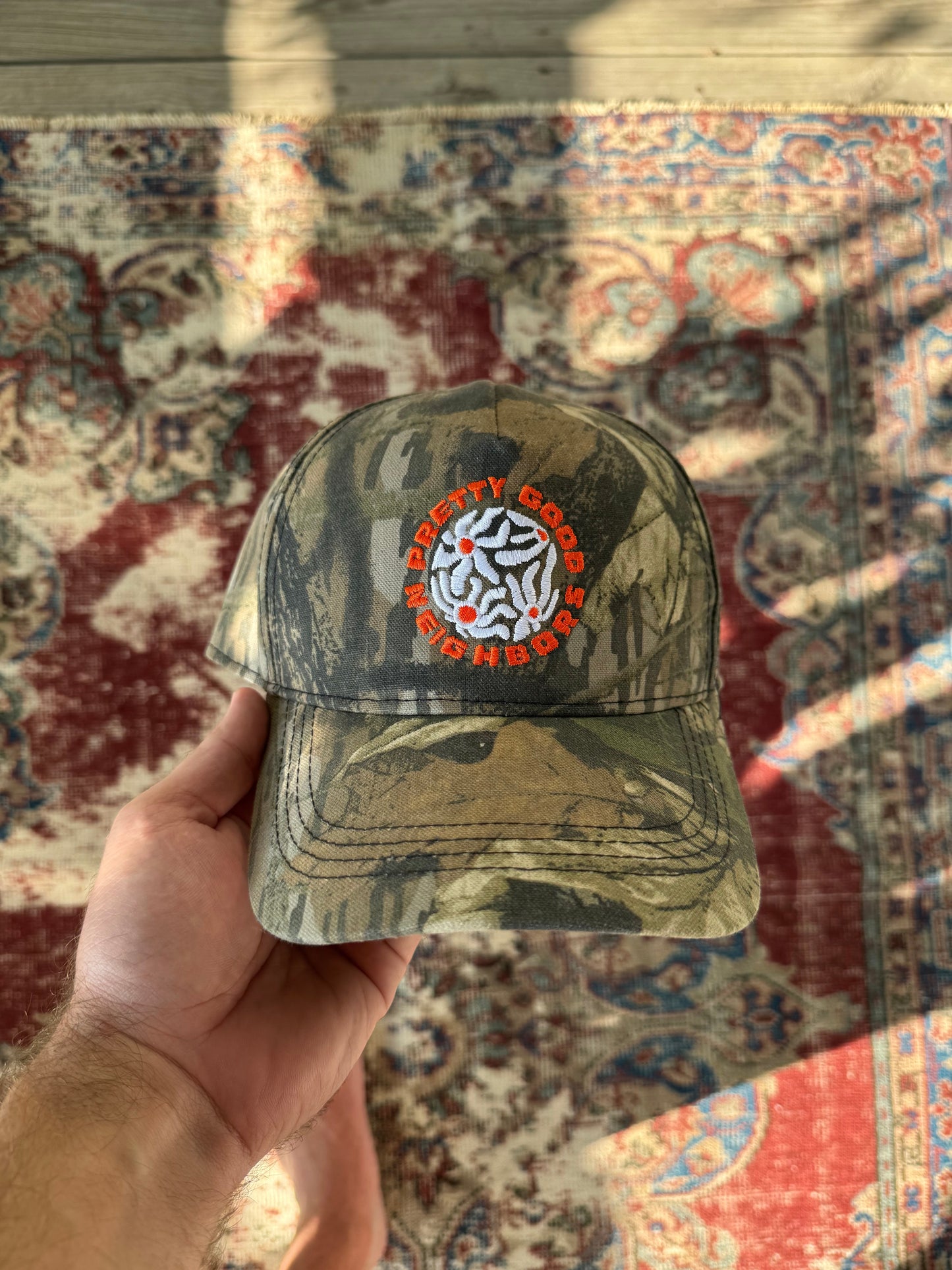 Camouflage-patterned cap with a logo held in front of a patterned rug