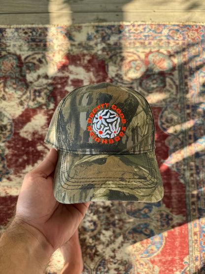 Camouflage-patterned cap with a logo held in front of a patterned rug