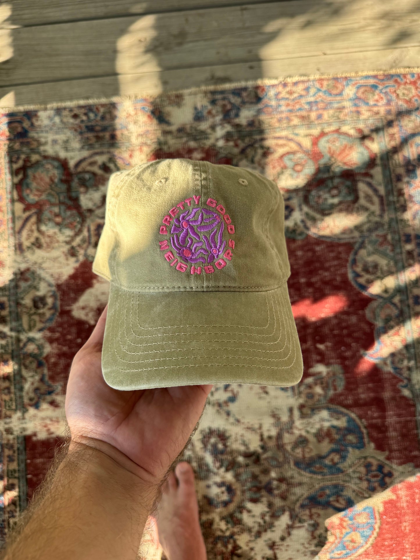 Khaki cap with a purple logo held by a hand on a patterned rug